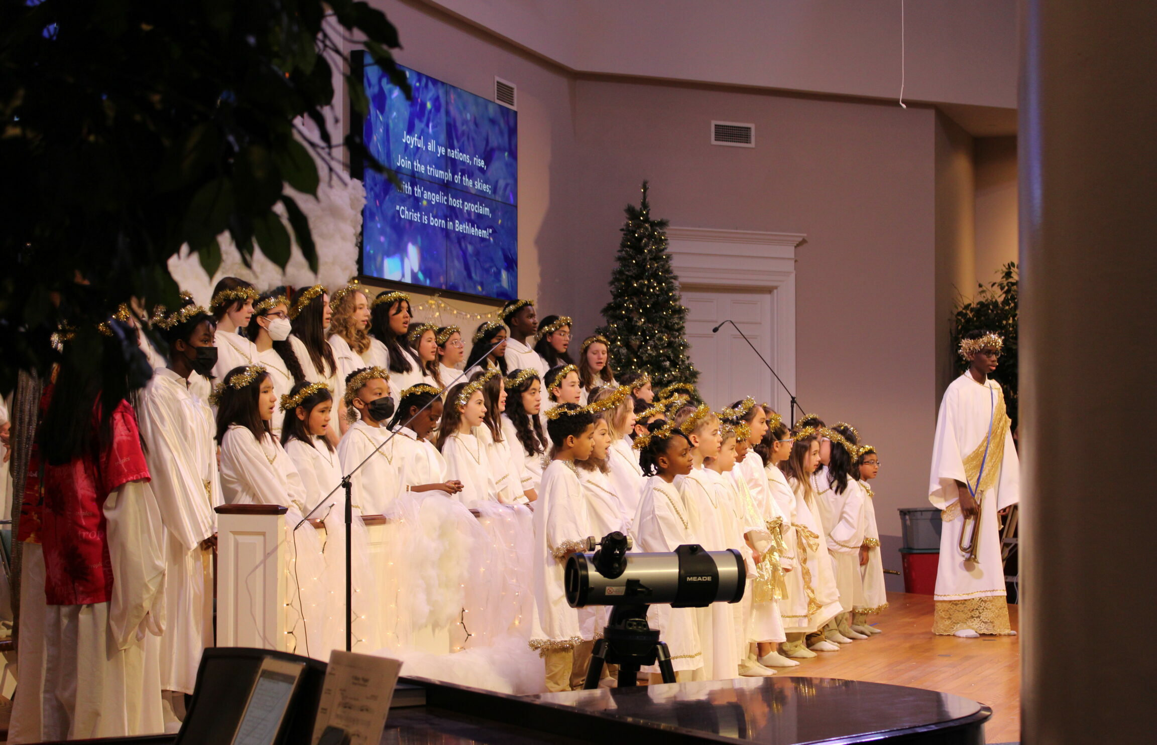 Photo of a children's choir, dressed in white robes and glittered halos, sing Christmas songs. One boy in the foreground holds a trumpet.