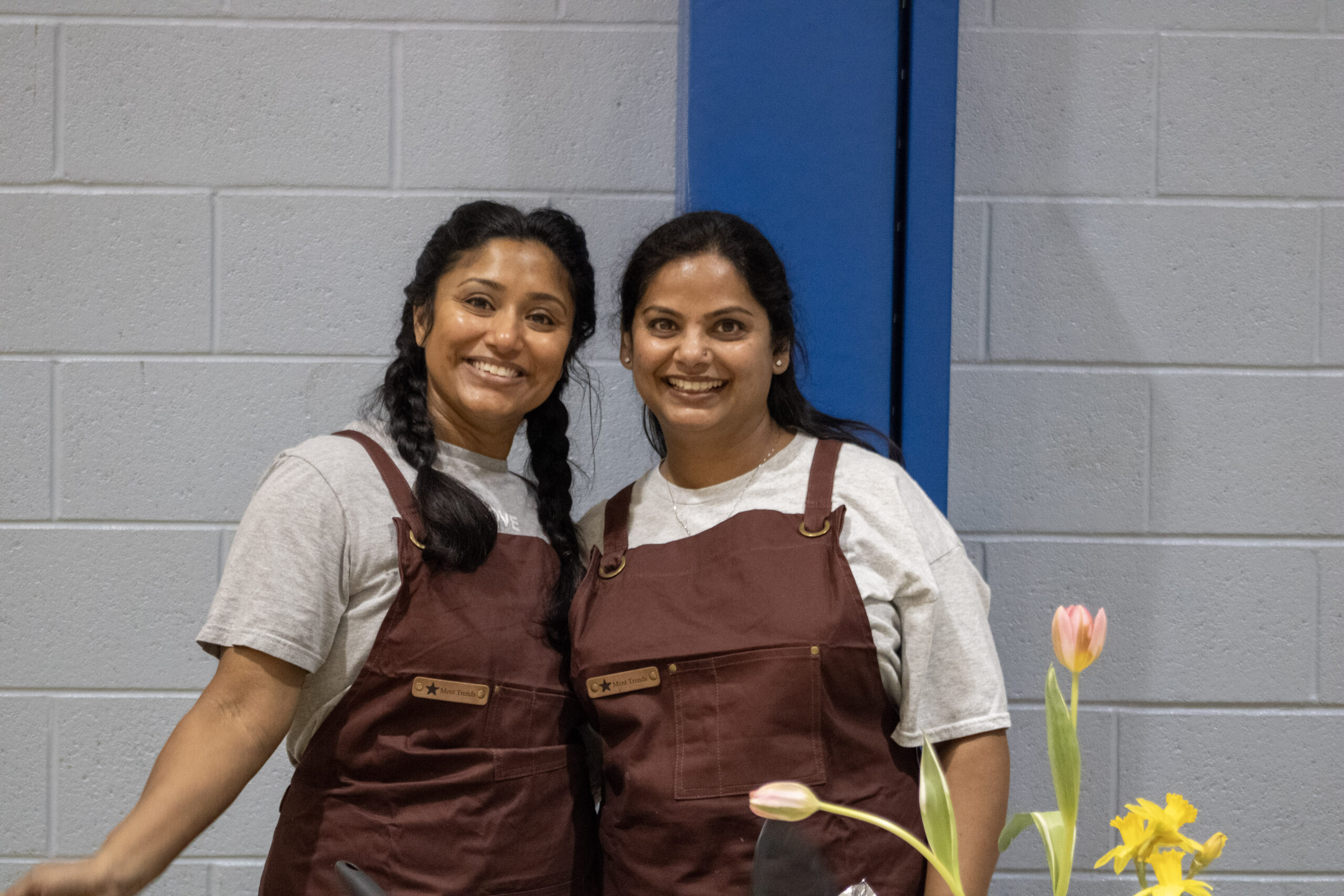 Two women pose together smiling. They are wearing matching gray shirts and brown overalls as outreach ministry volunteers. In the foreground is a bouquet of bright flowers.